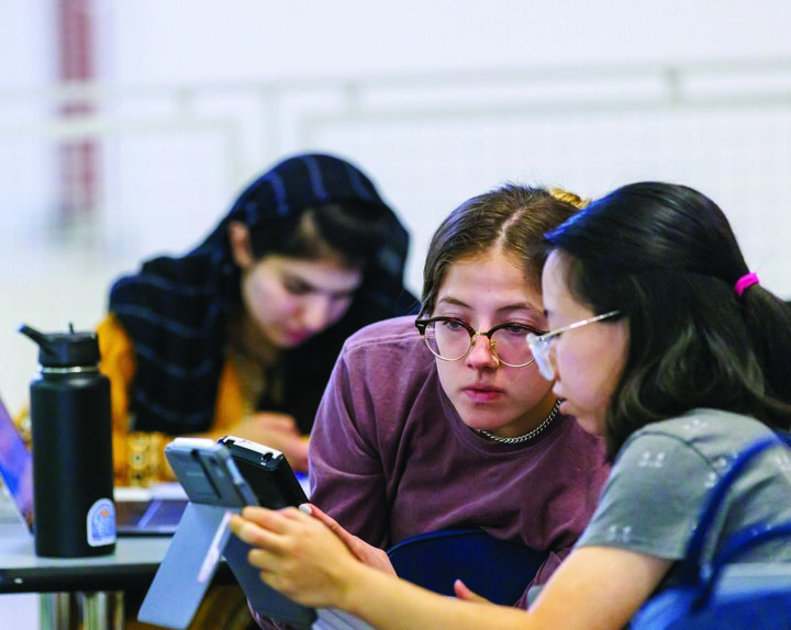 Two students using calculators with a third student on their phone in the background.
